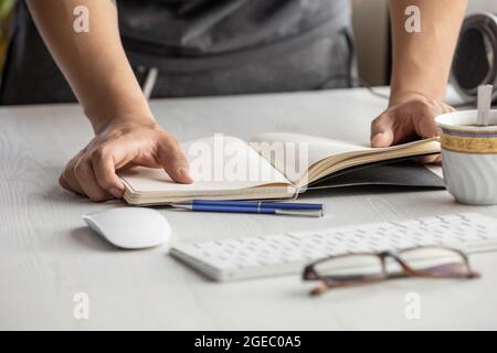 hands of a person holding an open and empty notebook, desk with glasses, pen, cup of coffee, computer mouse, keyboard, lifestyle with objects, studio Stock Photo