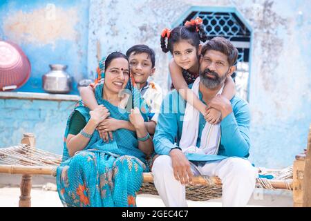 Portrait of happy rural indian family looking at camera while sitting on traditional bed at village home, little daughter and son hug father and mothe Stock Photo