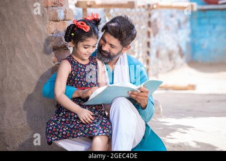 Happy rural indian father helping his daughter in studies, adorable little village girl child holding notebook studying outside home. Man teaching and