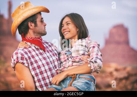 Lovers cowboy and cowgirl in love cute couple in USA backcountry landscape. Boyfriend wearing cowboy hat carrying Asian girlfriend seducing and Stock Photo