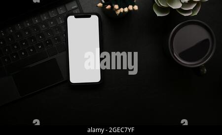 Modern workspace, smartphone white screen mockup on black magic keyboard, pencils, plant, coffee mug, black surface, top view, above Stock Photo