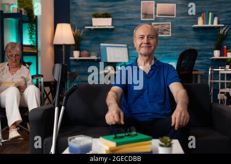 Elder mature man sitting on couch with crutches smiling looking at camera while retired woman in wheelchair with handicap focused on reading book in modern living room background Stock Photo