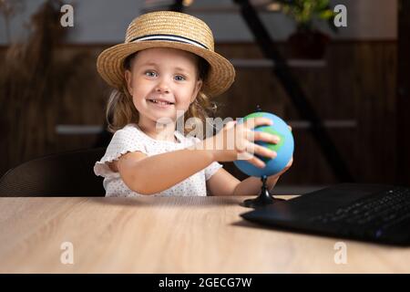 Caucasian cute little girl in a hat sits alone in the evening at a table with a laptop holding a globe in her hands. Stock Photo