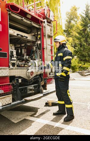 One young male firefighter dressed in uniform with protective helmet near fire engine. Side view Stock Photo