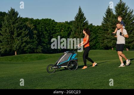 Family day. Happy young family with a child spend time together while walking with baby carriage on a green lawn in a city park on a weekend Stock Photo