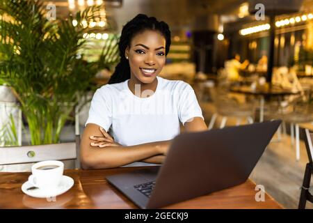 Portrait of young attractive African female blogger, typing a new challenging post during lunchtime at a cafe, with laptop and smart phone with copy s Stock Photo