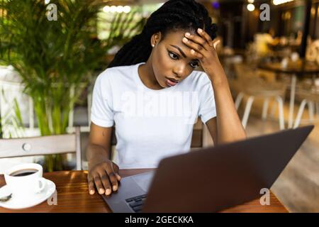 Pretty African woman sitting at table near laptop and looking at camera with tired face expression. Stock Photo