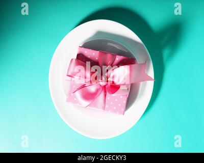 A gift box with a pink ribbon lies on the plates saucers on a mint-colored background Stock Photo
