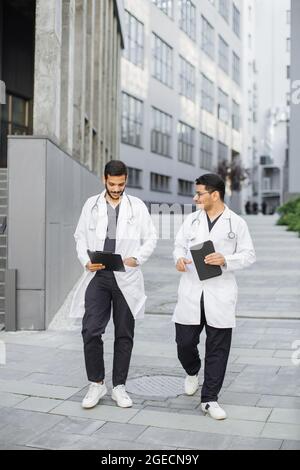 Two male Indian Arabian doctors in medical apparel, discussing patient's diagnosis, walking outside on the background of modern hospital Stock Photo
