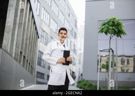 Male handsome Indian Asian doctor, wearing white coat, holding digital tablet in hands for working with patient's records, while standing outside mode Stock Photo