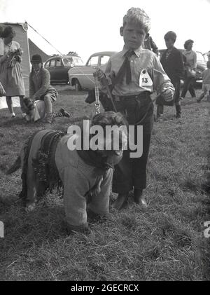 1950s, historical, a dog show with a difference, outside at a rural show, a fancy dress dog show, a small boy dressed as a cowboy with his pet dog also dressed in similar clothes, England, UK. Stock Photo