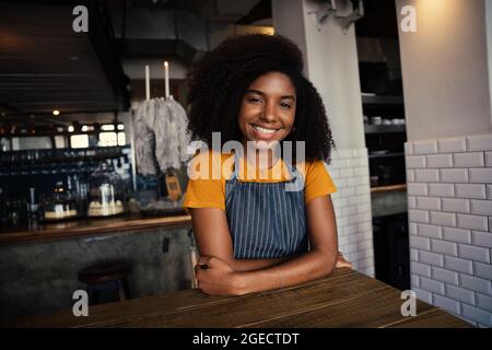 Beautiful mixed race female waitress wearing apron sitting at coffee table in funky cafe  Stock Photo