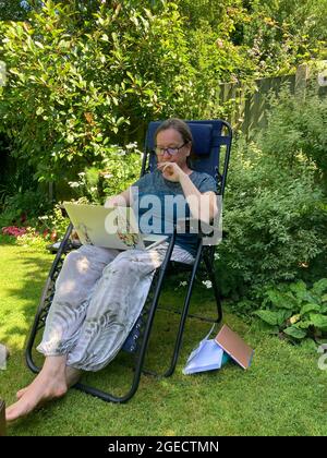 Woman using laptop computer, sitting outdoors in a lush green garden in the summer Stock Photo