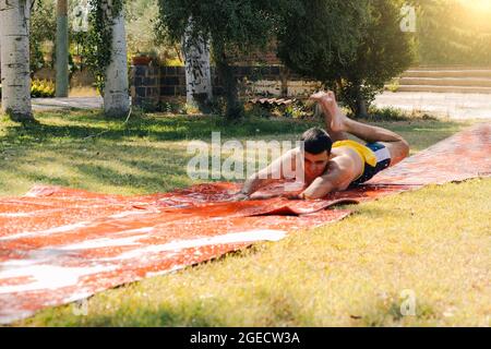 teenage boy in a bathing suit jumping down a soap and water slide or pool with his arms outstretched to slide down the garden and cool off on a hot su Stock Photo