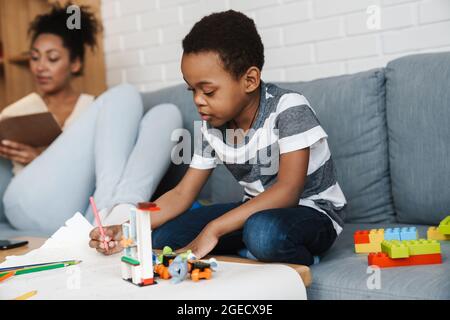 Black boy drawing and sitting on sofa while his mother reading book at home Stock Photo