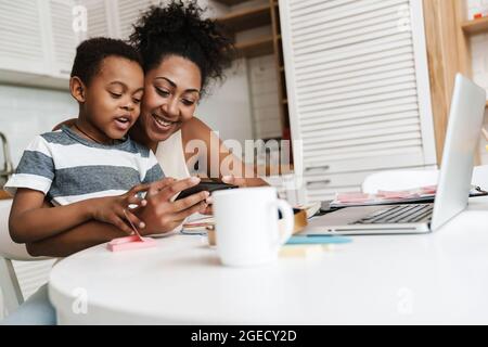 Black smiling mother and son using mobile phone and laptop while sitting together at home Stock Photo