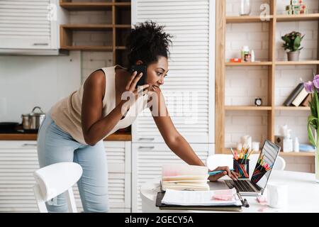 Black smiling woman talking on cellphone while working with laptop at home Stock Photo