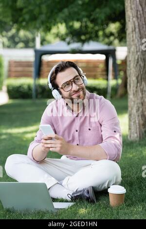 Dreamy businessman in headphones using gadgets near paper cup on grass Stock Photo