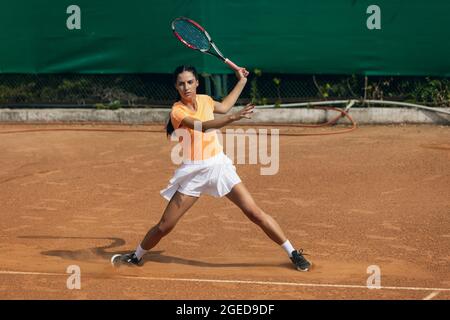 Beautiful young sportswoman, professional tennis player with racket practicing on clay tennis court in summer day. Stock Photo