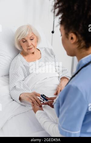 Elderly woman using digital oximeter near african american nurse in hospital ward Stock Photo