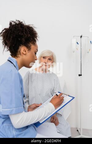 Senior patient looking at african american nurse writing on clipboard Stock Photo