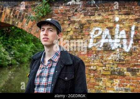 A young man standing by a bridge with the word pain spray painted onto the brickwork. The man looks deep in thought as he looks into the distance. Stock Photo