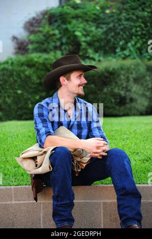 Young and handsome re-enactor sits waiting for his cue.  He is in a western theatre production and is wearing cowboy hat, jeans, gun belt and plaid sh Stock Photo