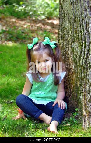 Pigtailed little girl enjoys going barefoot on a sunny afternoon in Tennessee.  She is sitting besides a tree in green grass. Stock Photo