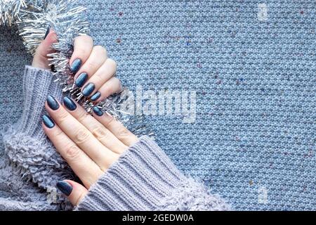 Female hands in gray knitted sweater with beautiful manicure - dark gray blue glittered nails on knitted background with silver Christmas tinsel garla Stock Photo