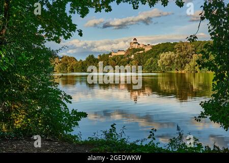 Castle above the river Vah. View of the right bank. Framed by tree leaves.  Reflection of beautiful clouds on the water surface. Trencin, Slovakia. Stock Photo