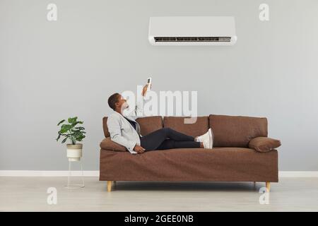 Black woman sitting on sofa, holding remote control and switching air conditioner Stock Photo