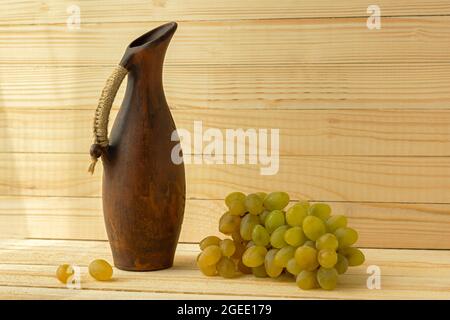 Grape jug on a wooden natural background. Rustic retro style. Antique clay jug with wine. Green juicy ripe grapes of the autumn harvest. the concept o Stock Photo