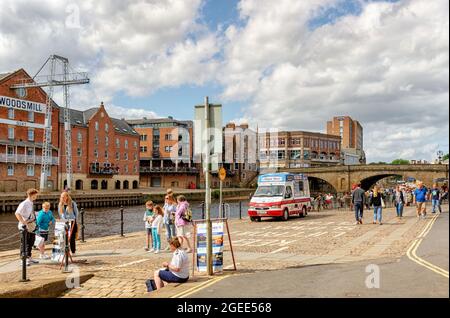 Riverside scene. A stone bridge spans a river with buildings on the other side. People are in the foreground and an ice-cream vendor is beside the riv Stock Photo