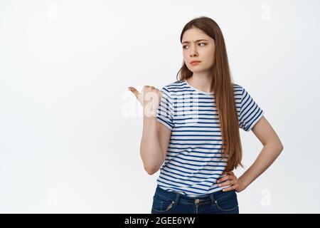 Image of skeptical young woman frowning, pointing and looking lef with doubtful, unimpressed face expression, standing over white background Stock Photo