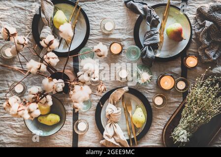 Fall or Autumn table styling for holiday dinner. Flat-lay of dinnerware with fruit, cutlery, candles and cotton flowers over beige tablecloth, top vie Stock Photo
