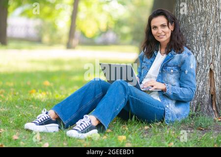 Portrait of a 40-years-old woman with laptop in city park during online working or distant online education. She looking at camera and smiling. Stock Photo