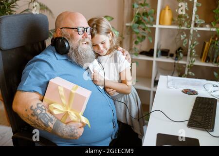 Happy man with gift box embraces blonde little girl holding greeting card in home office Stock Photo