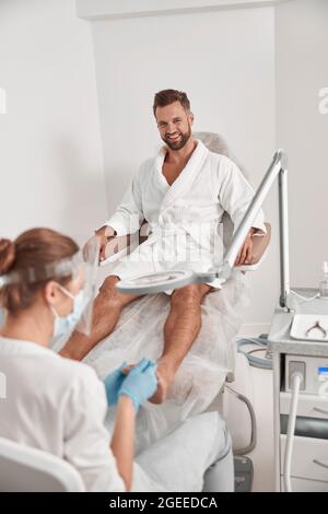 Smiling man in bathrobe sits in armchiar undergoing pedicure procedure in salon Stock Photo