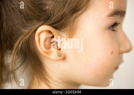 Ear piercing in a child - a girl shows an earring in her ear made of a medical alloy. White background, portrait of a girl with a mole on his cheek in Stock Photo