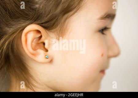 Ear piercing in a child - a girl shows an earring in her ear made of a medical alloy. White background, portrait of a girl with a mole on his cheek in Stock Photo