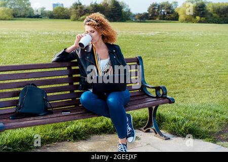 A woman in casual clothes drinking coffee from reusable cup and using a laptop outdoors sitting on the bench in the public park. Remote work Stock Photo