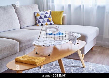 Washed towels on a table ironing board and an iron in a small living room with sofa, household routine time Stock Photo