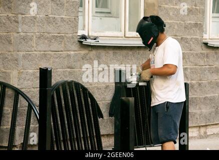 Male worker in protection mask sawing metal fence pieces with handheld circular saw, outdoors Stock Photo