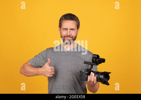 cheerful bearded man videographer making movie for vlog with camcorder showing thumb up, filming Stock Photo