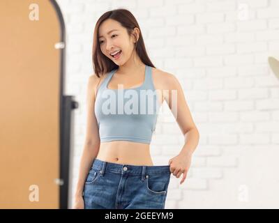 Happy young woman wearing large-size jeans , looks at her figure in front of the mirror. Stock Photo