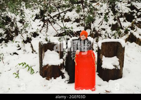 Caucasian boy sitting on a log seat in the snow holding a red toboggan Stock Photo