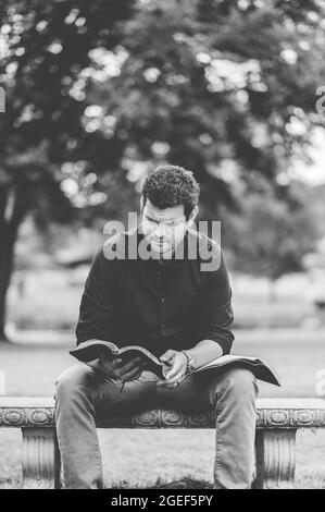 Vertical grayscale shot of a young Caucasian man reading the Bible while sitting on a bench Stock Photo