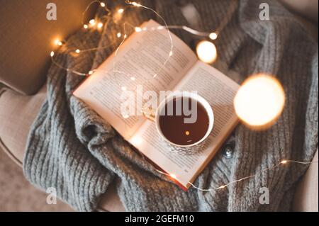 Cup of fresh black tea stay on open paper book with knit woolen sweater in cozy chair over glow Christmas lights closeup. Top view. Early morning brea Stock Photo