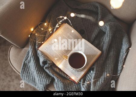 Cup of fresh black tea stay on open paper book with knit woolen sweater in cozy chair over glow Christmas lights closeup. Top view. Early morning brea Stock Photo