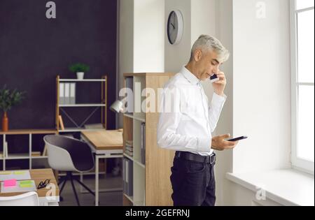 Serious mature businessman talking phone looking at digital tablet in office Stock Photo